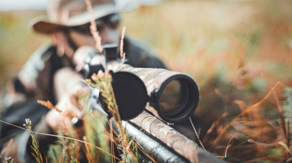 A hunter in a hat aiming a rifle in a grassy field, with the focus on the barrel of the gun.