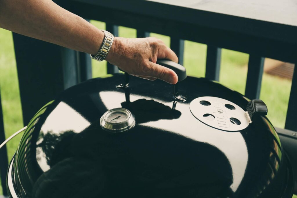 A person tending to a kettle grill, checking the temperature or adjusting the lid for a barbecue session outdoors.