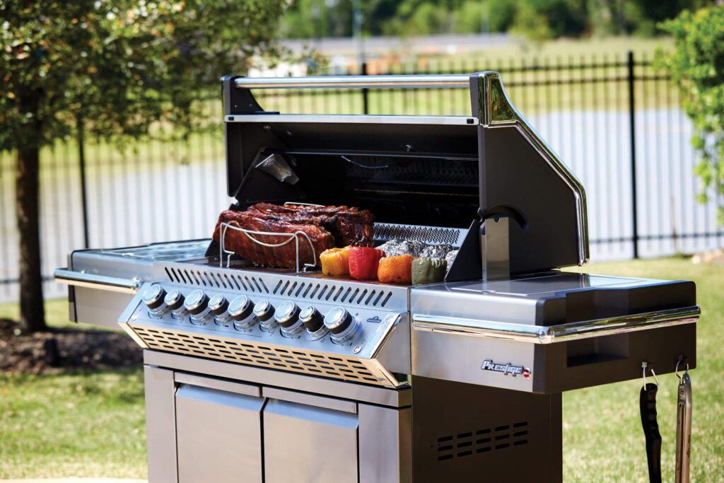 A shiny stainless steel gas grill cooking a large piece of meat and vegetables on a sunny day outdoors.