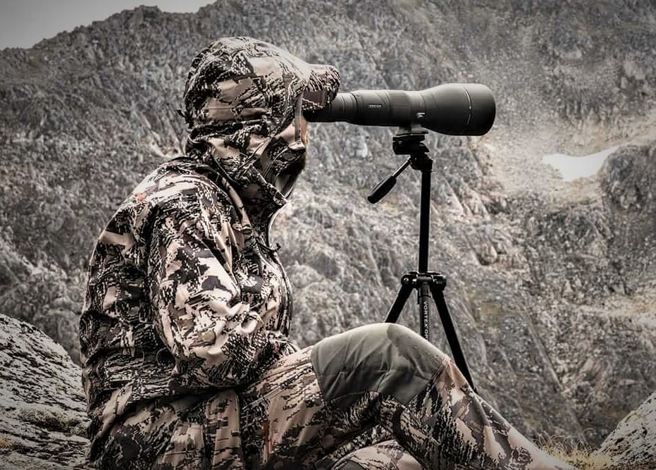 A camouflaged individual using Hunting Optics Brands long lens camera on a tripod to photograph or observe wildlife in a rugged mountainous terrain.